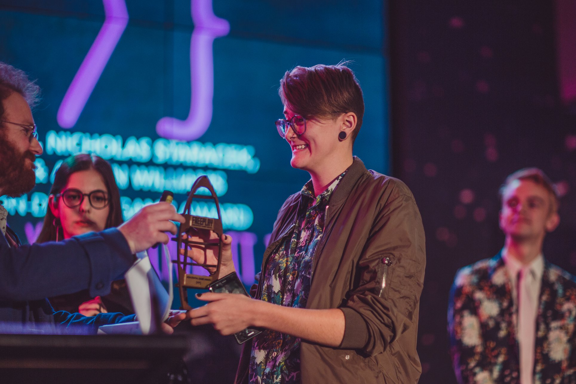 A smiling person with short hair accepts an award, shaped like a standing up boat, from another person with a beard. Behind them, two presenters look on, as a colourful projection lights up the background.